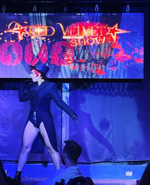 Cabaret performer with red hair in a top hat, tailcoat and sequined shorts striking a dramatic pose on a blue-lit nightclub stage in front of a neon show sign.