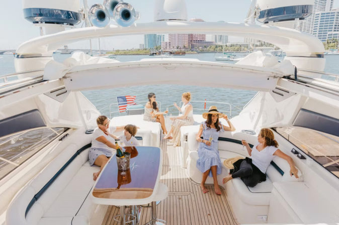 Friends relaxing on a luxury yacht in a sunny harbor, lounging on white seating, sipping champagne and cocktails with a city skyline and American flag in the background.