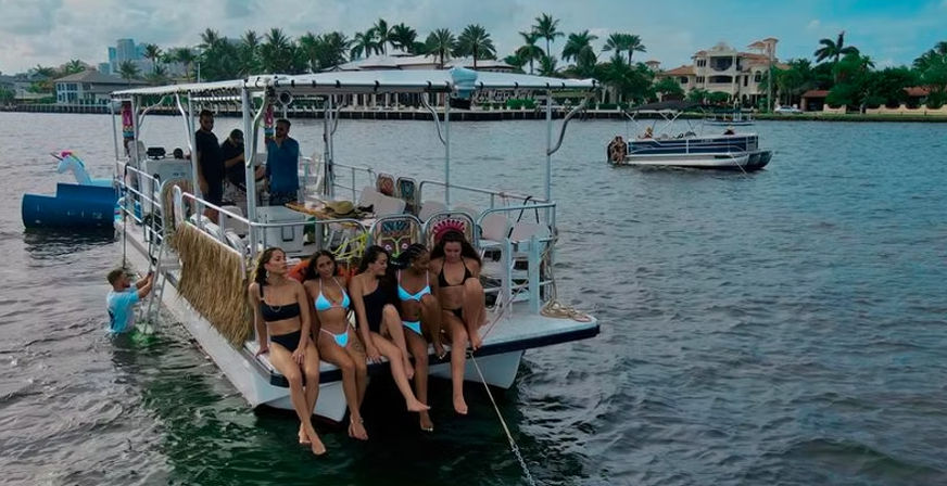 Group of women in bikinis sitting on the bow of a festive pontoon boat, friends and crew onboard, anchored on a palm‑lined coastal waterway with waterfront homes and another boat nearby.