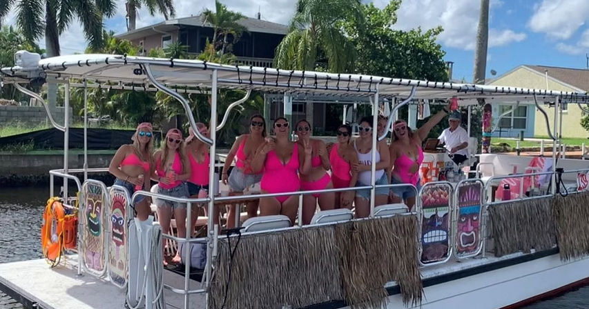 Group of women in bright pink swimsuits on a tiki‑decorated pontoon boat enjoying a sunny tropical canal cruise with palm trees and waterfront homes