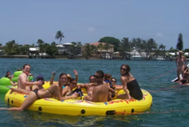 Group of adults relaxing on a bright yellow inflatable raft in a sunny tropical bay with clear blue water, palm-lined shoreline and waterfront homes in the background.