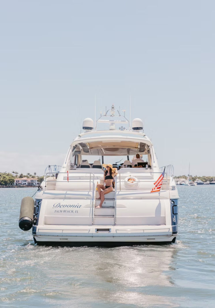 Sunny rear view of a white luxury yacht off the Florida coast with a woman in a black bikini and sunhat lounging on stern steps, an American flag fluttering and calm blue water with waterfront homes in the distance.