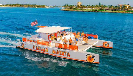 Orange-and-white party catamaran with an American flag and passengers enjoying a sunny cruise on clear turquoise coastal waters near a palm-lined shoreline
