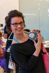 Smiling woman with glasses holding a colorful mosaic glass jar at an indoor craft fair or art workshop in a bright, casual setting.