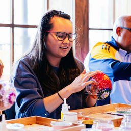Woman with glasses decorating a red beaded glass ornament at an indoor craft workshop by a sunlit window, with trays of beads, glue and tools on the table.