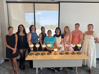 Ten women smiling and posing behind a table displaying nine colorful mosaic glass lamps in a bright conference room.