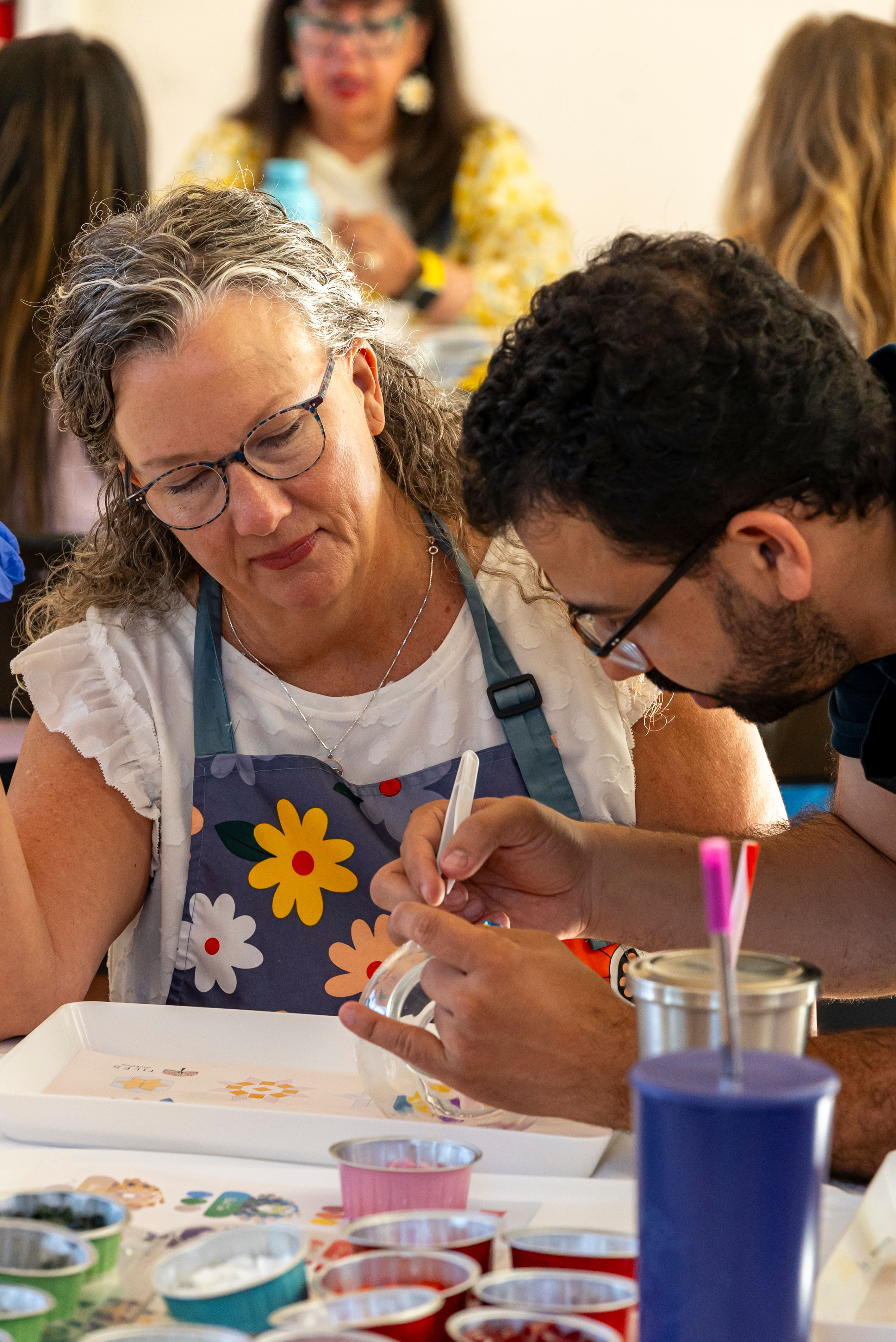 Two adults in a community art workshop paint a glass jar — a woman in a floral apron watches as an instructor guides detailed brushwork over a table of paint cups and tools.