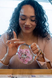 Close-up of a smiling woman at a DIY craft table decorating a clear glass ornament with pink beaded flower accents, handmade ornament crafting scene.