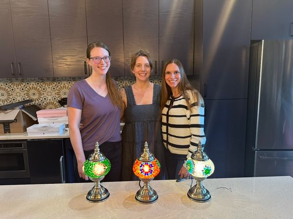 Three smiling people in a modern kitchen behind a quartz island showcasing three colorful Turkish mosaic table lamps (green, red/orange, white) with dark cabinets, patterned tile backsplash and stainless refrigerator.