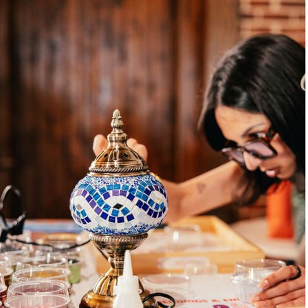 Ornate blue-and-white mosaic glass lamp on a brass stand surrounded by craft cups and glue, with a person leaning in to work in a cozy wooden craft workspace.