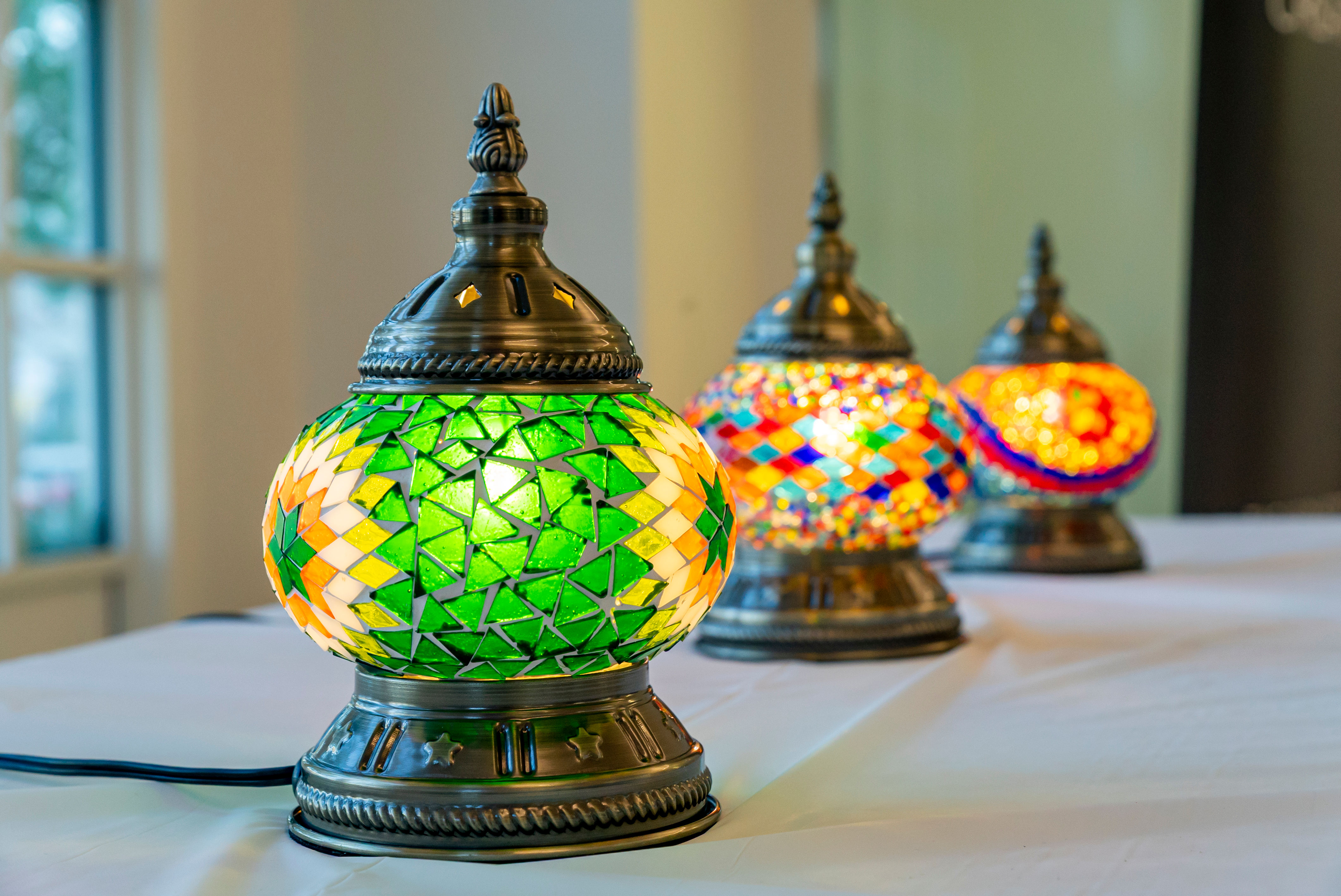 Vibrant Turkish-style mosaic glass table lamps on a white table, close-up of a glowing green mosaic lamp with ornate bronze base and blurred colorful lamps behind it.