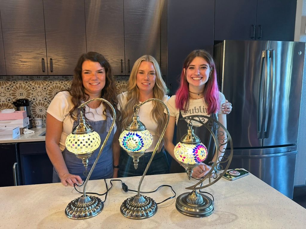 Three smiling women behind a kitchen island displaying three colorful handcrafted Turkish mosaic lamps on ornate bronze stands, with dark cabinets, patterned tile backsplash and stainless refrigerator in the background.