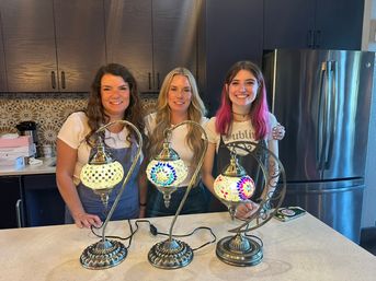 Three smiling women standing behind a kitchen island displaying three lit Turkish mosaic table lamps with colorful glass patterns, dark cabinets and stainless-steel refrigerator in a modern home kitchen.