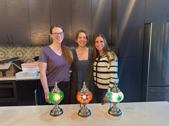 Three smiling women standing in a modern kitchen behind a white countertop displaying three lit colorful Turkish mosaic lamps; dark wood cabinets, patterned tile backsplash and stainless steel refrigerator visible.