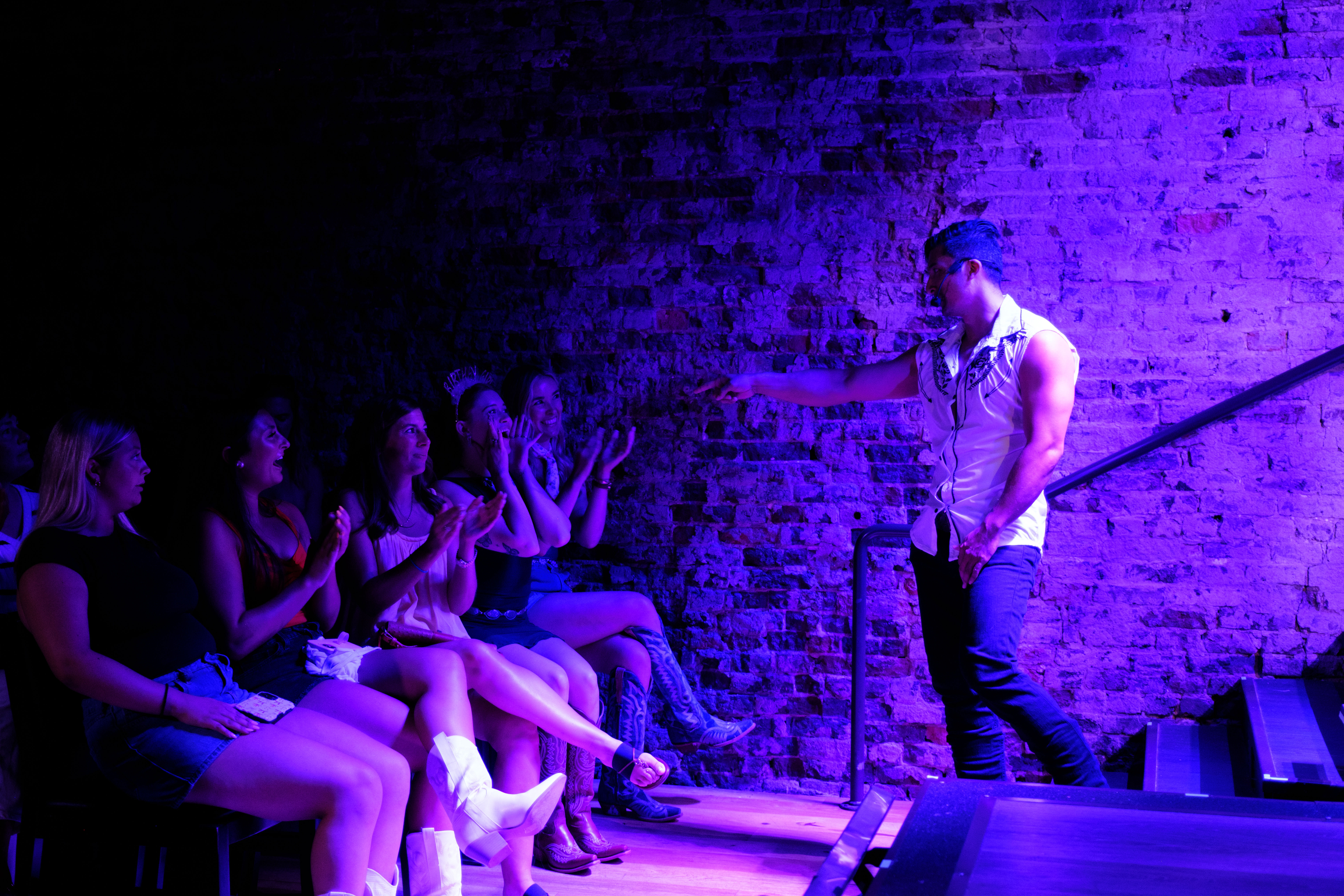 Purple-lit nightclub scene with a muscular male entertainer in a sleeveless shirt pointing from a brick-walled stage toward a row of cheering, clapping women in party attire, evoking a bachelorette-party vibe.