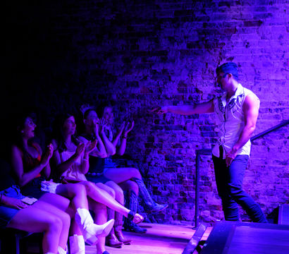 Purple-lit nightclub scene with a muscular male entertainer in a sleeveless shirt pointing from a brick-walled stage toward a row of cheering, clapping women in party attire, evoking a bachelorette-party vibe.