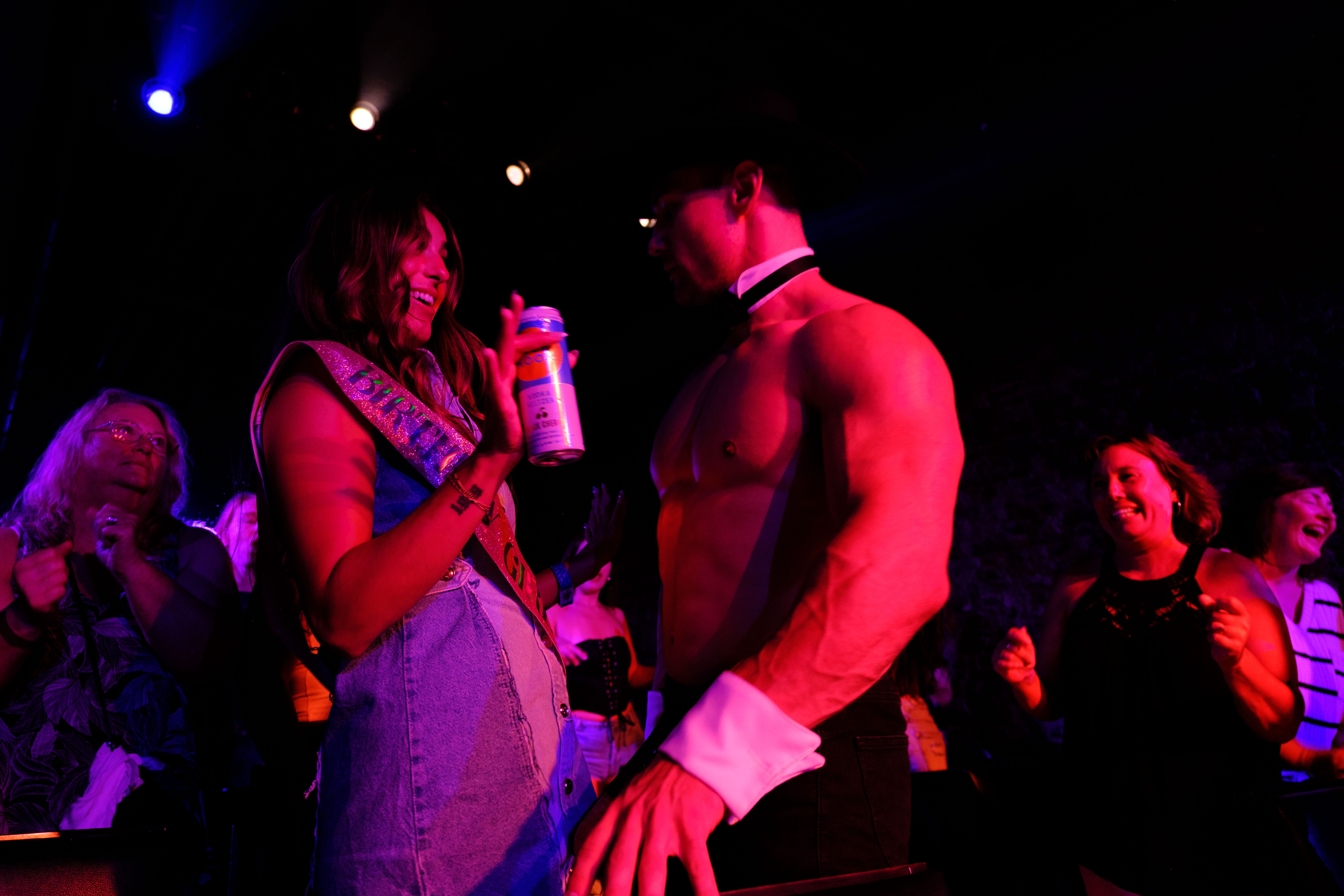 Nightclub bachelorette-style party: woman wearing a sash laughs and holds a drink while a shirtless male entertainer in collar and cuffs performs under purple-pink stage lights as cheering women watch.