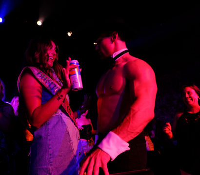 Nightclub bachelorette-style party: woman wearing a sash laughs and holds a drink while a shirtless male entertainer in collar and cuffs performs under purple-pink stage lights as cheering women watch.