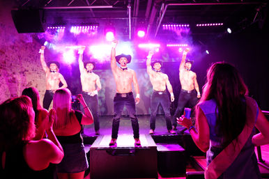 Shirtless male dancers in cowboy hats and bow ties strike a pose on a purple-lit nightclub stage while an excited audience films and cheers