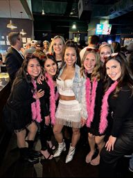 Group of smiling women posing at a crowded bar for a girls' night out, one wearing a sparkly fringe skirt and glitter boots while friends wear pink feather boas and novelty sunglasses.