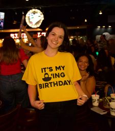 Smiling woman wearing a bright yellow 'It's my birthday' T-shirt posing in a dimly lit, crowded karaoke bar with neon signs and drinks in the background.
