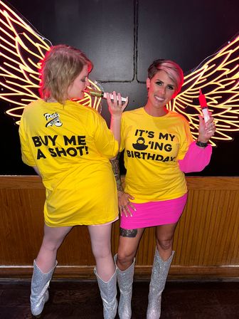 Two friends in bright yellow birthday shirts pose in front of neon angel wings at a bar, holding syringe-style shot dispensers and wearing silver glitter cowboy boots and a hot pink skirt.
