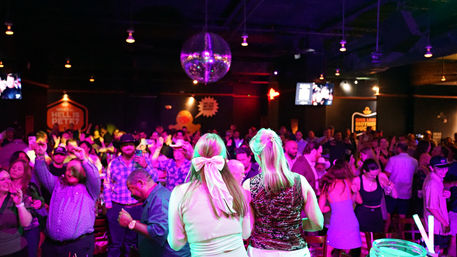 Back view of two women overlooking a packed nightclub dance floor with a glittering disco ball and purple neon lights.
