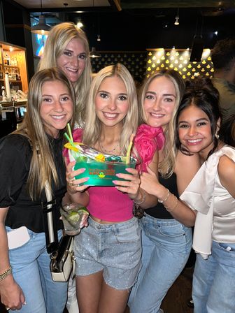Five smiling women posing in a lively indoor bar, holding a large blue-green shared cocktail bowl with straws and lime garnish during a fun night out