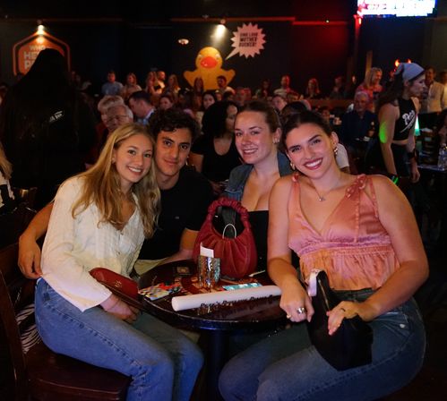 Four smiling friends posing at a crowded indoor bar table in a lively nightlife scene, drinks on the table and a large yellow rubber duck mural on the back wall