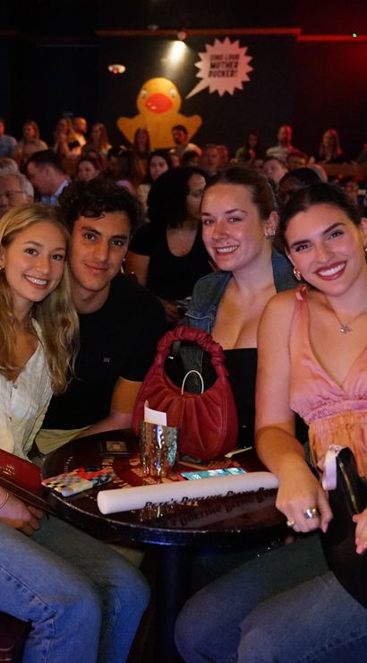 Four smiling friends posing at a crowded indoor bar table in a lively nightlife scene, drinks on the table and a large yellow rubber duck mural on the back wall