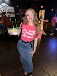Smiling woman in a pink graphic tee and denim skirt holding a large multi-straw fishbowl cocktail inside a dimly lit bar with wooden floors and other patrons in the background.