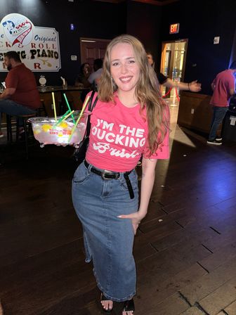 Smiling woman in a pink graphic tee and denim skirt holding a large multi-straw fishbowl cocktail inside a dimly lit bar with wooden floors and other patrons in the background.