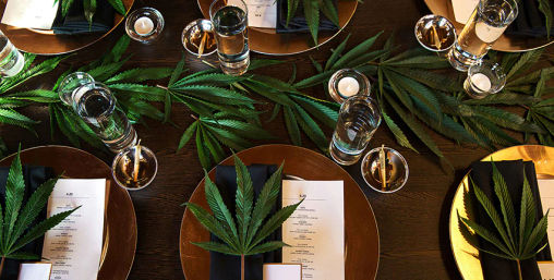 Overhead view of an elegant cannabis-themed dinner table with gold charger plates, black napkins, printed menus, large cannabis leaves as place-setting decor, glassware and tealight candles on a dark wooden table.