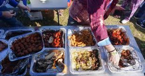 Outdoor buffet in a park with aluminum trays of fried and grilled dishes (wings, fries, fritters) and a gloved hand serving.