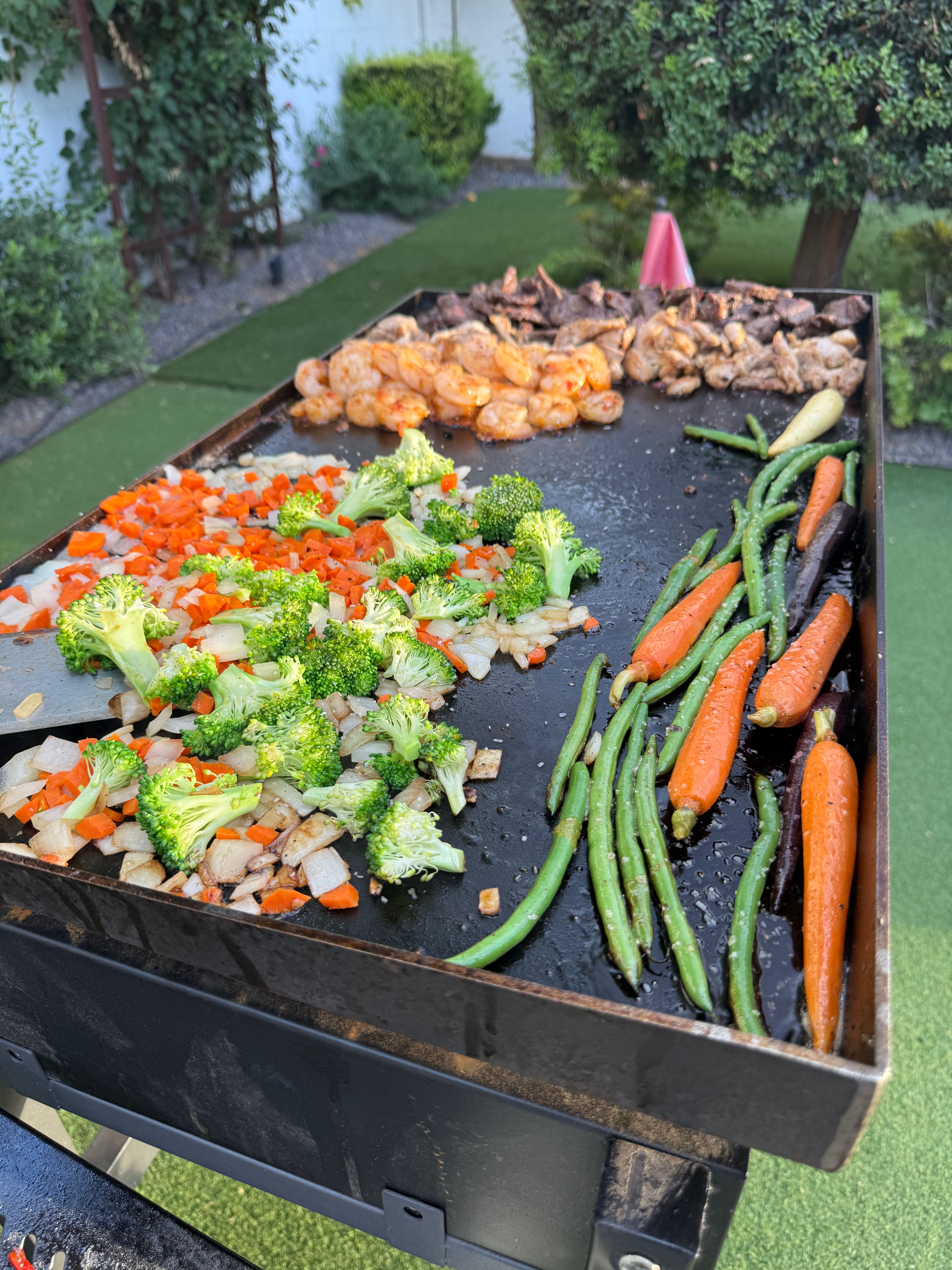 Backyard flat-top griddle sizzling with broccoli, diced onions and carrots, green beans, baby carrots and seasoned shrimp in a garden setting.
