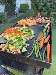 Backyard flat-top griddle sizzling with broccoli, diced onions and carrots, green beans, baby carrots and seasoned shrimp in a garden setting.
