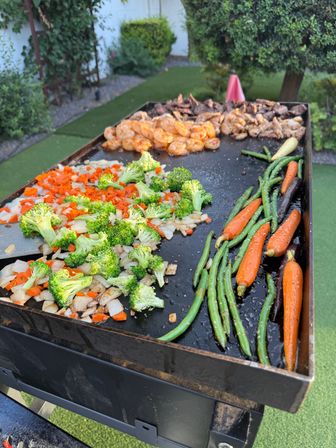 Backyard flat-top griddle sizzling with broccoli, diced onions and carrots, green beans, baby carrots and seasoned shrimp in a garden setting.
