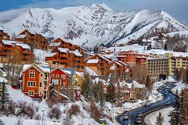 Cozy colorful chalets and lodges in a snowy mountain ski town, framed by evergreen trees, a winding roadway, and towering snow‑capped peaks.