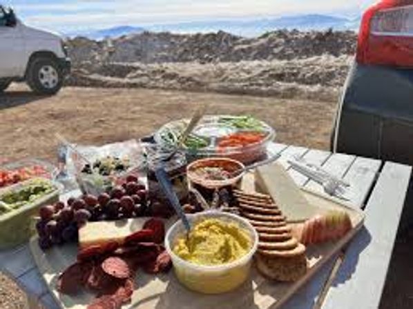 Charcuterie-style mountain picnic: cheeses, crackers, cured meats, grapes, hummus and veggie dips on a picnic table by parked cars at a snowy overlook.
