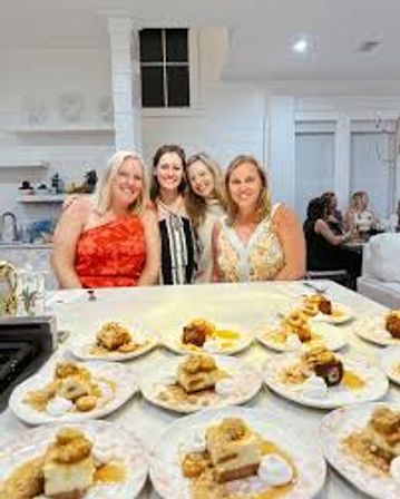 Four smiling women posing behind a bright modern white kitchen island filled with plated desserts — friends enjoying a dessert-tasting gathering.