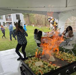 Backyard tented party with a caterer’s flaming griddle cooking mixed vegetables while a photographer snaps photos; guests seated at a decorated table with black, white and gold balloon garland near a suburban house.
