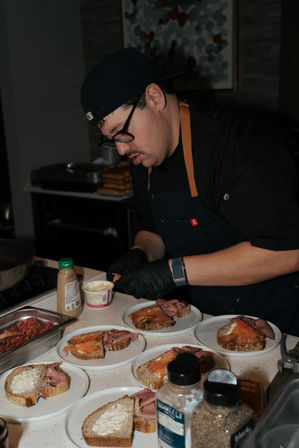 Chef in black apron and gloves carefully plating open-faced sourdough sandwiches topped with smoked salmon and roast beef on plates in a busy commercial kitchen.