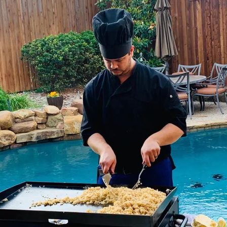 Backyard chef in black uniform flipping fried rice on a large flat-top griddle next to a turquoise pool, with stone landscaping, wooden fence and patio seating in the background.