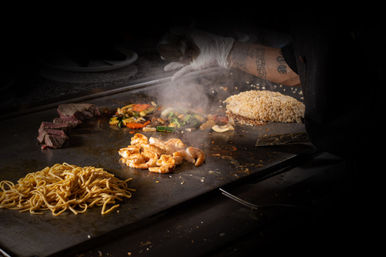 Hibachi chef's gloved hand sprinkling seasoning over sizzling shrimp on a flat-top griddle, surrounded by fried rice, lo mein noodles, seared steak slices and mixed vegetables with steam rising.