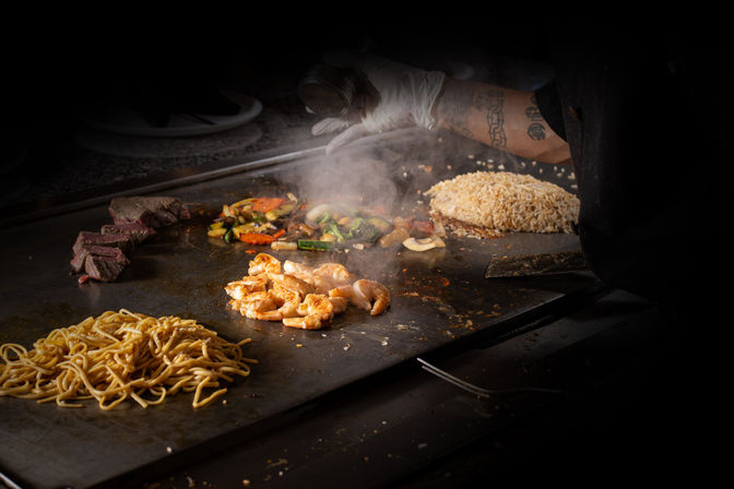 Hibachi chef's gloved hand sprinkling seasoning over sizzling shrimp on a flat-top griddle, surrounded by fried rice, lo mein noodles, seared steak slices and mixed vegetables with steam rising.