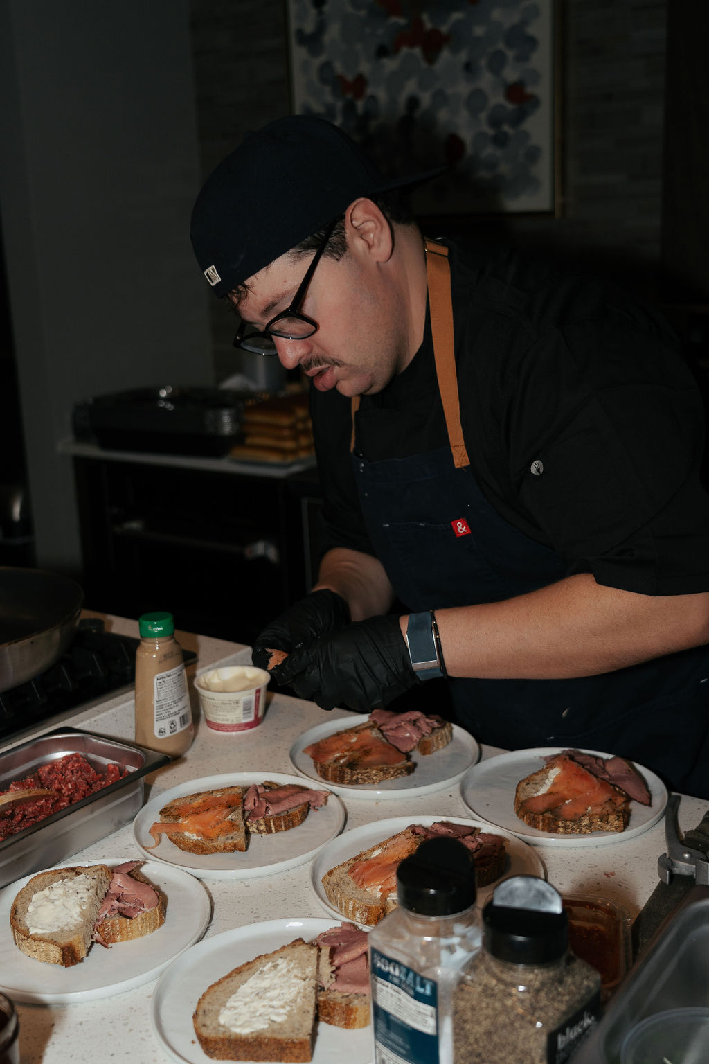 Chef in a busy restaurant kitchen plating open-faced smoked salmon and roast beef sandwiches on rustic sourdough, with condiments and spice jars on the counter