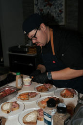 Chef in a busy restaurant kitchen plating open-faced smoked salmon and roast beef sandwiches on rustic sourdough, with condiments and spice jars on the counter