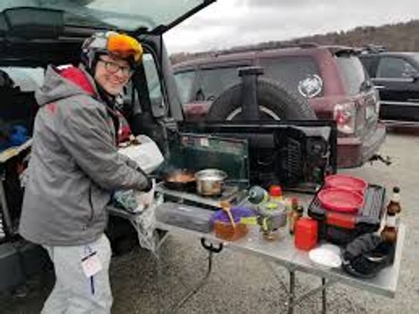 Smiling person tailgate-cooking at an SUV in a parking lot — portable stove, pots, coolers and utensils on a folding tailgate table on a cloudy day.