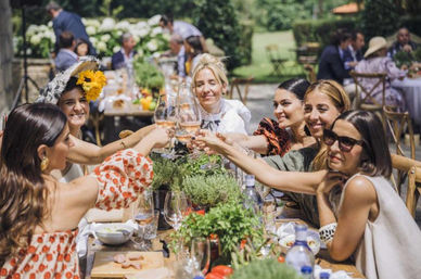 Smiling women toasting with wine glasses around a rustic wooden table at a sunny outdoor garden brunch, wearing summer dresses and hats with fresh herbs and shared plates on the table.