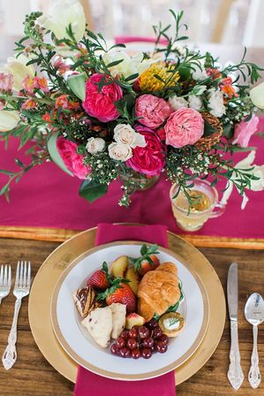 Elegant wedding tablescape with a vibrant pink-and-white floral centerpiece on a magenta runner and a gold charger holding a croissant, strawberries, grapes and assorted pastries